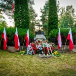Pomnik otoczony flagami Polski, kwiaty i znicze na trawie. W tle drzewa i krzewy tworzą zielone tło. Kolory dominujące to czerwień, biel i zieleń.