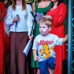 Na zdjęciu grupa osób na scenie, w jasnych ubraniach. Kobiety trzymają mikrofony, młody chłopiec z przodu ma białą koszulkę z Garfieldem. Tło jest zielone, sugerując plener.