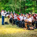 Orkiestra dęta w mundurach gra na zewnątrz. W tle są muzycy i zielone drzewa. Flagą Polski jest widoczna po prawej stronie.