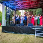 Grupa ludzi na scenie na tle drzew, w kolorowych strojach, z mikrofonami. Niebiesko-czerwone światła. Obok scena z mikrofonem.