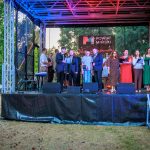 Grupa osób stoi na scenie pod zadaszeniem, w tle drzewa. Dominują kolory zielony i czerwony. Scena ma napisy Powiat Miński.