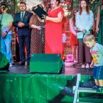 Grupa ludzi stojąca na scenie, kobieta w czerwonej sukni przemawia. Tło zielone z drzewami i napisem Powiat Miński. Na dole sceny chłopiec schodzi po schodkach.