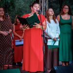 Na scenie stoi kobieta w czerwonej sukni, przemawiając do mikrofonu. W tle inne osoby, drzewa i metalowa konstrukcja. Tło jest zielone, obrazem sceny plenerowej.