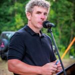 Mężczyzna w czarnej koszulce mówi do mikrofonu na tle zielonego lasu. Trzyma niebieski przedmiot. Obok znajduje się pojazd.