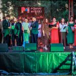 Grupa ludzi na scenie śpiewa do mikrofonów. Są ubrani kolorowo, na tle drzew i baneru „Powiat Miński”. Na scenie widać zielone światła.