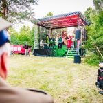 Plenerowa scena w parku z występem zespołu muzycznego, kolorowe stroje artystów. Publiczność siedzi na trawie obok stołu. Zielone drzewa w tle.