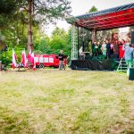 Na scenie w parku ludzie w różnorodnych strojach występują. Obok sceny stoją biało-czerwone flagi i wóz strażacki. Tło stanowią zielone drzewa.
