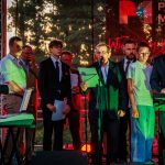 Grupa osób stoi na scenie w eleganckich ubraniach z mikrofonami i papierami. Tło wypełnione jest drzewami i światłami. Dominuje zielony i czerwony kolor oświetlenia.
