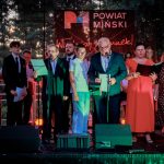 Grupa ludzi stoi na scenie z mikrofonami, w tle napis Powiat Miński. Tło zielone z drzewami, oświetlenie w czerwono-zielonym kolorze. Ubrani są elegancko, w garnitury i sukienki.