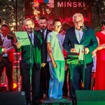 Na scenie stoi grupa osób z mikrofonami, ubranych w kolorowe stroje. Tło jest ciemne, z zielonymi i czerwonymi światłami. Na plakacie widoczny jest napis „MIŃSKI”.