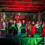 Grupa ludzi śpiewa na scenie w kolorowym świetle, ubrani w eleganckie stroje. Tło stanowi ciemny las z przezroczystym logo Powiat Miński. Na scenie widać również sprzęt muzyczny.