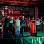 Grupa osób stoi na scenie, śpiewając do mikrofonów. Tło to baner z napisem POWIAT MIŃSKI oraz drzewa. Scena oświetlona jest ciepłym, czerwonym światłem.