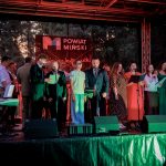 Grupa osób śpiewa na scenie z napisem Powiat Miński. Tło jest ciemne, z leśnym krajobrazem. Uczestnicy mają mikrofony i nuty.