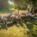 Orkiestra dęta w białych uniformach gra na trawniku w słoneczny dzień. Tło lasu, scena i polskie flagi. Różnorodne instrumenty.