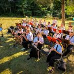 Orkiestra dęta w mundurach gra na świeżym powietrzu. Sceneria to zielona łąka z drzewami w tle. Instrumenty błyszczą, a dyrygent prowadzi z nutami na stojaku.