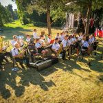 Orkiestra dęta na świeżym powietrzu gra koncert. Muzycy w białych koszulach siedzą na trawie. W tle drzewa i scena.