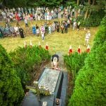 Ceremonia upamiętnienia przy grobie otoczonym zielenią, z tłumem ludzi w tle. Widać białe i czerwone flagi. Dominują kolory zielony i brązowy.