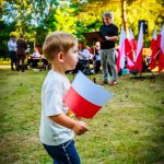 Na zdjęciu chłopiec trzyma polską flagę. W tle grupa ludzi oraz drzewa w parku. Sceneria jest jasna i słoneczna.