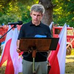 Mężczyzna w czarnej koszuli przemawia przy mównicy na tle lasu. Obok niego są biało-czerwone flagi. W tle widoczny czerwony pojazd strażacki.