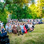 Grupa ludzi siedzi na krzesłach na trawie w zielonym parku. W tle drzewa i jasne, słoneczne światło. Ubrania różnobarwne, miejsce przypomina wydarzenie plenerowe.