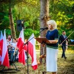 Kobieta przemawiająca przy mikrofonie w lesie, otoczona polskimi flagami. Tło pełne zielonych drzew. Uroczysta atmosfera.