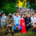 Grupa ludzi siedzi na zewnątrz na krzesłach, niektórzy trzymają polskie flagi. Na pierwszym planie człowiek w mundurze, obok kobieta w czerwonej sukni. Tło to zieleń drzew i liczny tłum w dalszym planie.