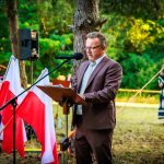 Mężczyzna w garniturze przemawia na świeżym powietrzu obok drzew i polskich flag. Tło to zielone drzewa, a za nim widoczni ludzie. Atmosfera wydarzenia jest formalna.