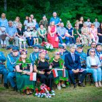 Grupa ludzi siedzi na zewnątrz, niektórzy w historycznych mundurach. W tle drzewa. Widać polskie flagi i kwiaty.