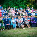 Na zdjęciu widoczna jest uroczystość na świeżym powietrzu. Ludzie siedzą na krzesłach w parku, trzymając polskie flagi. W tle widać drzewa i zielone liście.