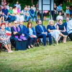 Grupa ludzi siedzi na krzesłach w plenerze podczas ceremonii. Tło jest zielone, z drzewami i tłumem na stojąco. Uczestnicy trzymają flagi Polski.