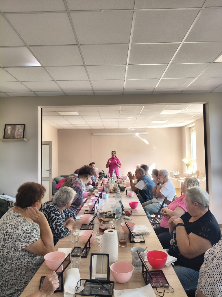 W jasnym pomieszczeniu, przy długim stole, siedzi grupa ludzi z różowymi miskami. Przód sali zajmuje osoba w różowym stroju. Na stole butelki wody i papiery.