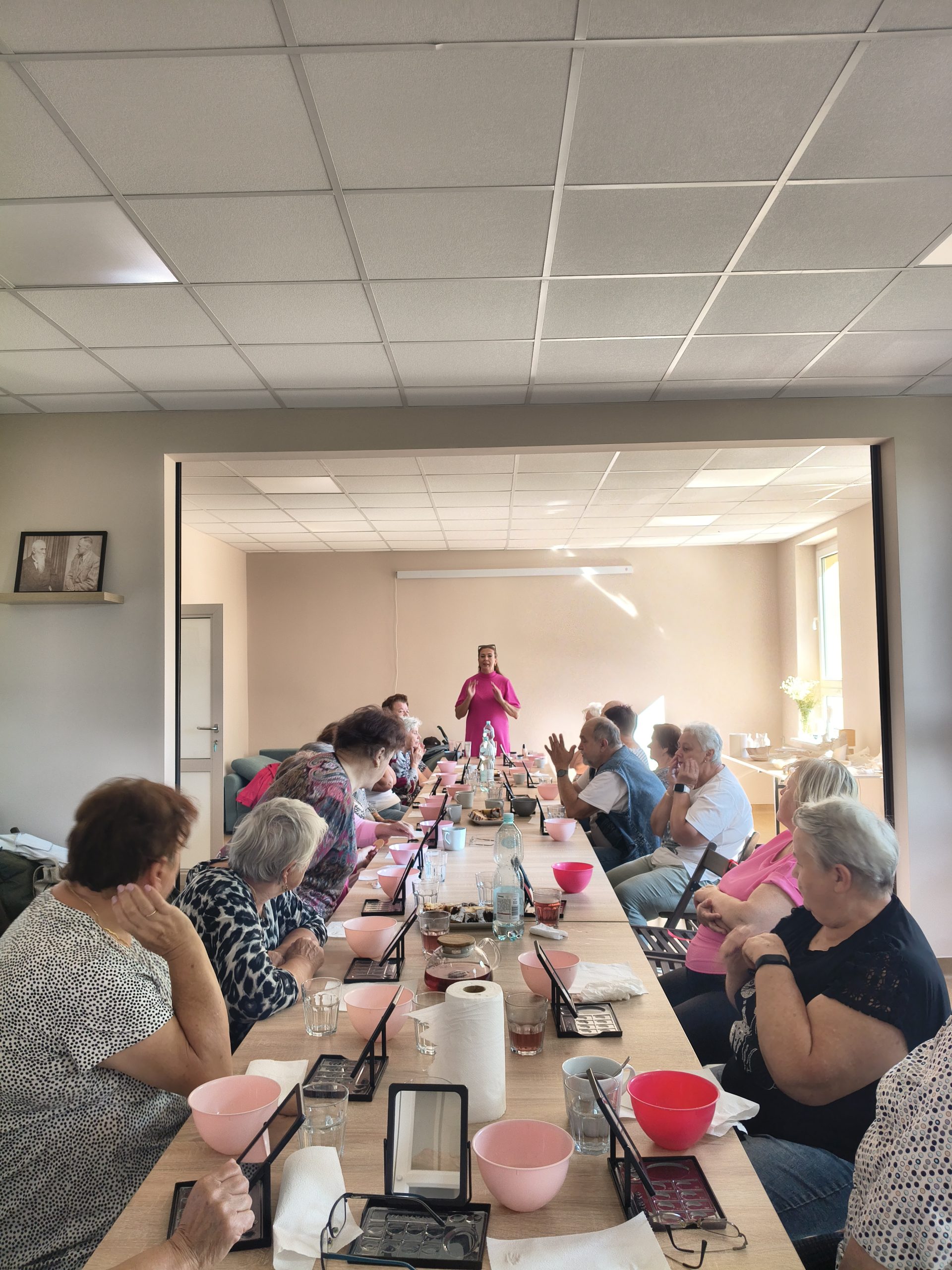 W jasnym pomieszczeniu, przy długim stole, siedzi grupa ludzi z różowymi miskami. Przód sali zajmuje osoba w różowym stroju. Na stole butelki wody i papiery.