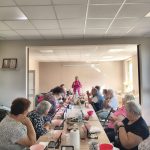 Grupa osób siedzi przy długim stole w jasnym pokoju. Na stole są różowe miski i czarne kosmetyczki. Osoba na końcu stołu prowadzi prezentację.