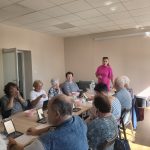 Na zdjęciu grupa seniorów siedzi przy stole, używając lusterek. Kobieta w różowej bluzce prowadzi zajęcia. Pokój jest jasny, z beżowymi ścianami.