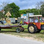 Traktor z czerwoną przyczepą ozdobioną kwiatami stoi na tle zielonych drzew i nieba. Przyczepa przystrojona kolorowymi wstążkami i sianem. Tło stanowią zaparkowane auta i tablice reklamowe.