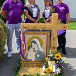 Grupa czterech osób w fioletowych koszulkach stoi za ozdobnym wieńcem z wizerunkiem Madonny z Dzieciątkiem, otoczonym kwiatami. W tle biały budynek i zieleń. Ozdoba składa się z kłosów zboża i kolorowych kwiatów.