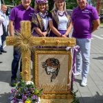 Grupa ludzi w fioletowych strojach stoi obok dzieła z kłosów i kwiatów. W tle drzewa i niebo. Centralny element kompozycji to religijny wizerunek z kłosów.