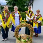 Cztery kobiety w ludowych strojach z kolorowymi chustami stoją za dożynkowym wieńcem z pszenicy i owoców. Tło to zielone krzewy i biała ściana budynku. Wieńce zdobią żółte i pomarańczowe akcenty.