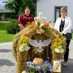 Dekoracja z kłosów zboża z białym ptakiem pośrodku, ozdobiona kwiatami i owocami. Przód domu w tle, zielone drzewa. Dwoje ludzi stoi obok, pogoda słoneczna.