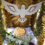 Chleb z napisem DOŻYNKI DOBRET 2026 na białej tkaninie. Obok winogrona i kwiaty. W tle dekoracja z zbożem i białym gołębiem.