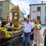 Grupa ludzi w białych koszulach przed wozem ozdobionym słomianą dekoracją i słonecznikami. W tle dwa budynki. Niebieskie niebo z chmurami.