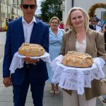 Dwie osoby trzymają dekoracyjne chleby na białych serwetach. Tło stanowią ludzie i budynek w jasnych kolorach. Atmosfera jest uroczysta.