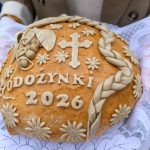 Na zdjęciu widać bochen chleba ozdobiony napisem DOŻYNKI 2026, kwiatami i krzyżem. Tło jest białe, koronkowe. Bochen trzymany jest w rękach na obrusem.