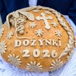 Chleb dożynkowy z napisem Dożynki 2026, ozdobiony warkoczem i kwiatami na jasnym tle z jasnoniebieską tkaniną.