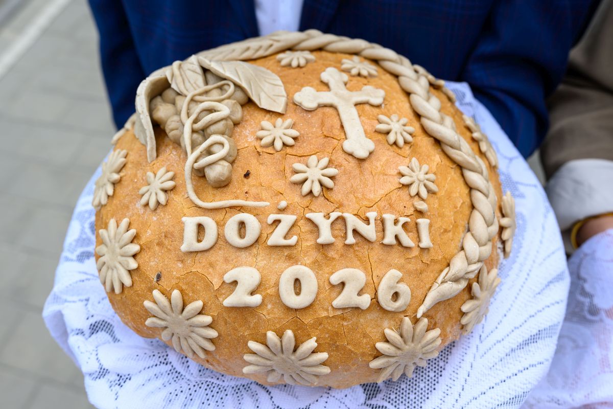 Chleb dożynkowy z napisem Dożynki 2026, ozdobiony warkoczem i kwiatami na jasnym tle z jasnoniebieską tkaniną.