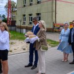 Grupa ludzi stoi przed budynkiem, niektórzy w białych koszulach, z chlebem na tacy. Tło jest zielone z drzewami. Budynek w kolorze kremowym z oknami.