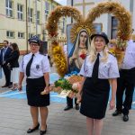 Na zdjęciu dwie kobiety w białych koszulach i ciemnych spódnicach w procesji z wizerunkiem Maryi. Tło stanowi budynek szkoły.