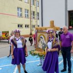 Święto religijne z osobami w fioletowych strojach i wieńcem. Tło to budynek w żółci i szarości. Na chodniku niebieskie płytki.
