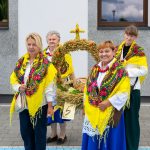 Na zdjęciu cztery kobiety w kolorowych chustach trzymają dożynkowy wieniec zbożowy. Tło to biało-szary budynek. Kobiety ubrane są w biel i odcienie niebieskiego.