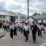 Parada strażaków w mundurach maszerujących obok budynku z flagą Polski. W tle pochmurne niebo. Widoczne instrumenty muzyczne.